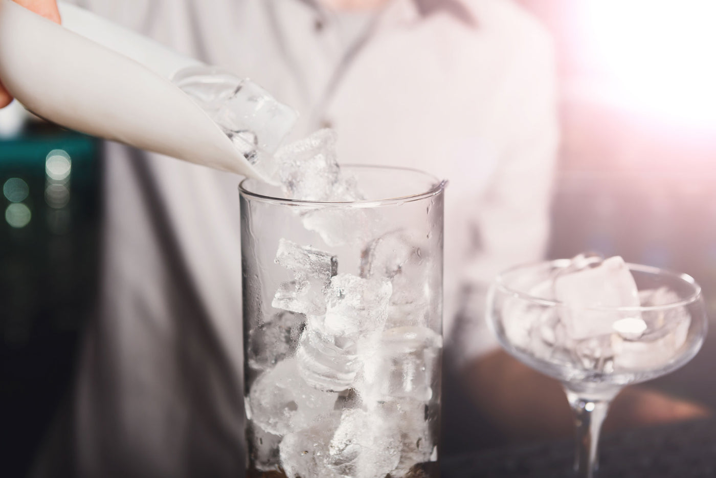 Clear glass highball tumbler being filled with ice cubes from a white scoop held by a bartender, with a coupe glass of ice on a dark bar counter in the background.